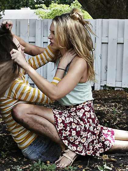 Two playful girls enjoy mud wrestling,  one of them blows a hung